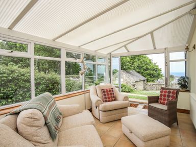 A sunroom with a beige sofa and armchairs with cushions and a plaid blanket and a floor lamp and open glass doors showing a garden at Fron Fawr in Nefyn