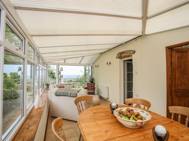 A sunroom with a wooden dining table and chairs a sofa and large windows overlooking greenery at Fron Fawr in Nefyn