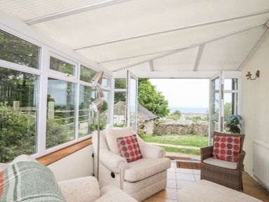A sunroom with beige armchairs a wicker chair and an open door leading outside at Fron Fawr in Nefyn
