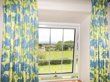 A window with blue floral curtains looking out onto a green field with houses and trees at Fron Fawr in Nefyn