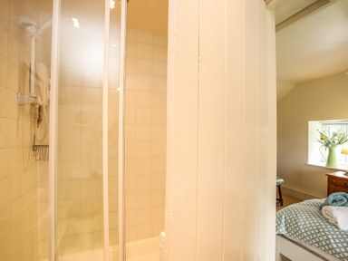 A shower with glass doors and a bathroom wall next to a bedroom with a bed a window and a vase of flowers at Fron Fawr in Nefyn