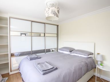 A bedroom with a bed covered in gray bedding and two folded towels on top a wardrobe with sliding doors and mirrors and a bedside table with a lamp and a small plant at The Gables in Trearddur Bay
