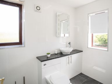A bathroom with a white toilet and countertop sink under a mirror and two windows at The Gables in Trearddur Bay