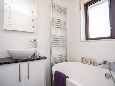 A bathroom with a white basin on a black countertop a towel rack a bathtub with a purple towel and a window at The Gables in Trearddur Bay