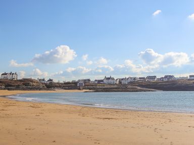 A sandy beach with calm water and houses on rocky shore under blue sky at The Gables in Trearddur Bay