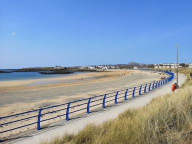 A sandy beach with a blue railing along a promenade and houses in the background at The Gables in Trearddur Bay