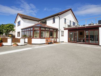 The exterior of a two story white house with a brown roof a sunroom with wooden framed windows a wooden bench and a patio area at The Gables in Trearddur Bay