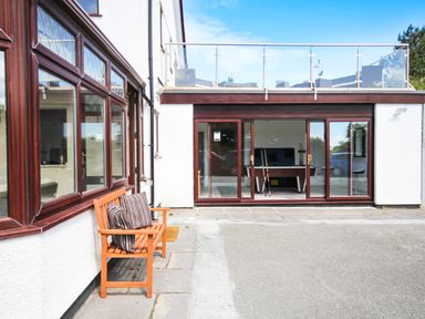 An outdoor seating area with a wooden bench and cushions next to a building with large windows and glass sliding doors showing a pool table inside at The Gables in Trearddur Bay