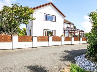 A detached two-story house with white walls brown roof and windows a glass conservatory a wooden fence and a gravel driveway at The Gables in Trearddur Bay