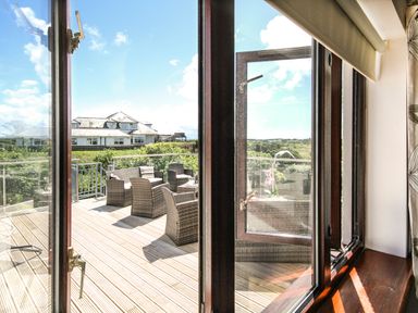 A balcony with wicker chairs and a table outside a glass door overlooking greenery and a house in the distance at The Gables in Trearddur Bay