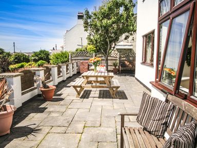 A patio with a wooden bench and striped cushions a wooden picnic table with flowers in a vase potted plants and a tree at The Gables in Trearddur Bay