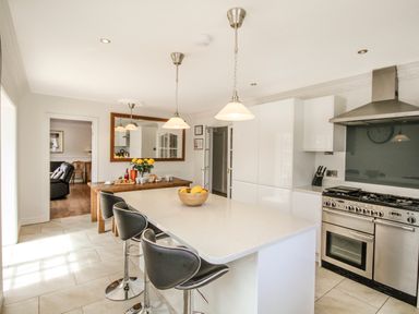 A kitchen with a large island with bar stools a wooden table with flowers and food and a stainless steel oven at The Gables in Trearddur Bay