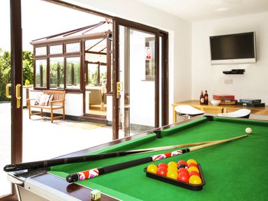 A room with a pool table with balls and cues and a TV on the wall and a door leading to an outdoor seating area at The Gables in Trearddur Bay