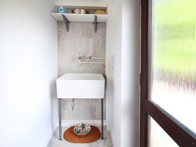 A utility room with a white sink and two taps above it a metal bowl on a mat below the sink and a shelf with items above at The Gables in Trearddur Bay