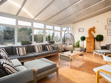 A sunroom with a sectional sofa, coffee table, hanging chair, wooden chest, and potted plants at The Gables in Trearddur Bay