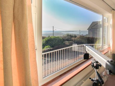 A window with beige curtains overlooking a garden and a distant view of water with a telescope on a tripod in the foreground at Garnedd Lwyd in Llanfair
