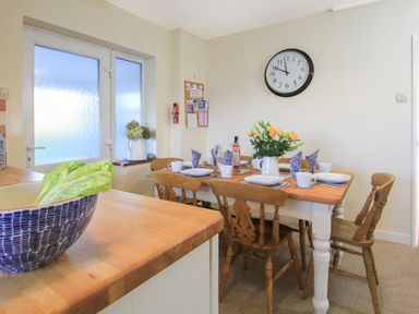 A dining area with a wooden table set with plates cups and napkins wooden chairs a large clock on the wall a bowl with lettuce on the counter and flowers on the table at Garnedd Lwyd in Llanfair