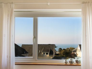 A window with white curtains overlooking rooftops and the sea with four decorative pitchers on the windowsill at Garnedd Lwyd in Llanfair
