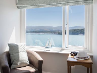A room with a chair, pillow, small wooden table with a hat and binoculars by a window overlooking water and hills at Glan Dwr Bach in Glyn Garth