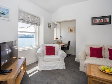 A living room with white sofas and red cushions a wooden table with fruit a TV on a wooden stand and a desk with a chair near windows at Glan Dwr Bach in Glyn Garth