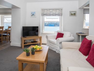 A living room with white armchairs and sofa with red cushions a wooden coffee table with plants and fruit a TV on a wooden stand and a dining area visible through an open doorway at Glan Dwr Bach in Glyn Garth
