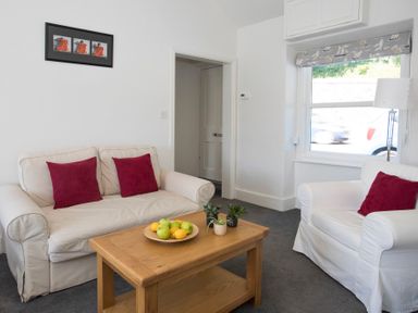 A living room with white sofas with red cushions a wooden coffee table with fruit and plants a window with patterned blind and a lamp at Glan Dwr Bach in Glyn Garth