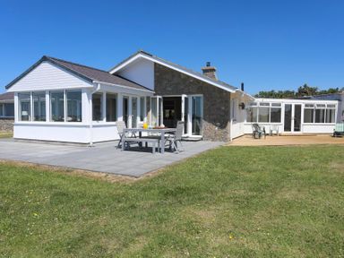 A white and stone house with a patio table and chairs on a paved area and plastic chairs on a wooden deck in a grassy yard at Glanrhyd Uchaf in Llanfair yn Neubwll