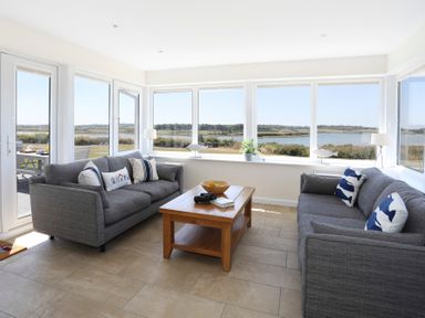 A living room with two grey sofas and a wooden coffee table by large windows overlooking a river at Glanrhyd Uchaf in Llanfair yn Neubwll