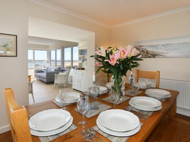 A dining room with a table set for six with plates glasses and flowers overlooking a living room with large windows and a view of water at Glanrhyd Uchaf in Llanfair yn Neubwll