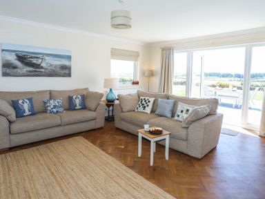 A living room with two beige sofas cushions a small table with cake and mug a lamp and large glass doors leading outside at Glanrhyd Uchaf in Llanfair yn Neubwll