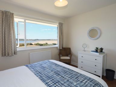A bedroom with a bed a blue blanket a wicker chair a white dresser with plants and a mirror and a window with a view at Glanrhyd Uchaf in Llanfair yn Neubwll