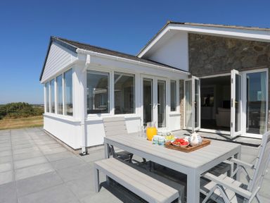 An outdoor patio with a dining table set and fruit jug near a house with large windows and open doors at Glanrhyd Uchaf in Llanfair yn Neubwll
