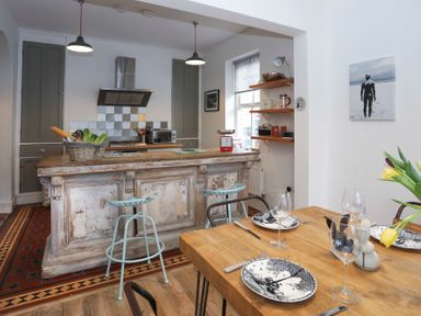 A kitchen with a wooden island and bar stools next to a dining table set with plates and glasses at Gwalia Stores in Beaumaris