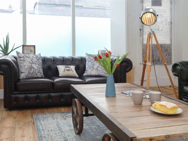 A living room with a leather sofa, a wooden coffee table with a vase of red tulips, two mugs, a plate with cake, and a tripod floor lamp at Gwalia Stores in Beaumaris