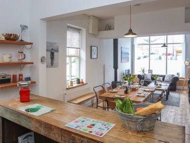 A kitchen and dining area with wooden table set for meals and a basket of vegetables on the counter at Gwalia Stores in Beaumaris