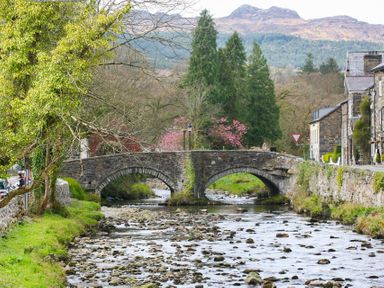 A stone bridge over a shallow river with trees and stone buildings on either side at Gwyndy Bach in Beddgelert