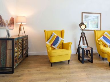 A living room with yellow armchairs a wooden floor lamp and a wooden sideboard at Gwyndy Bach in Beddgelert