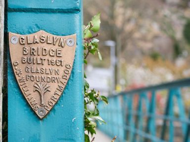 A metal plaque on a teal painted bridge railing with foliage and a blurred background at Gwyndy Bach in Beddgelert
