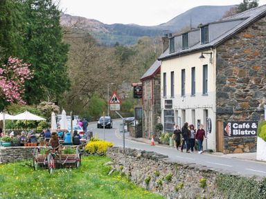 A street with a cafe and bistro sign stone buildings outdoor seating with umbrellas and people walking on the sidewalk at Gwyndy Bach in Beddgelert