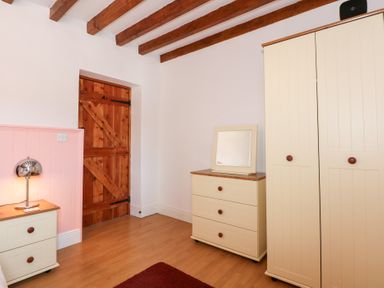 A room with wood ceiling beams a wooden door a cream wardrobe a cream chest of drawers with a mirror on top and a cream bedside table with a lamp at Hafod in Pontllyfni