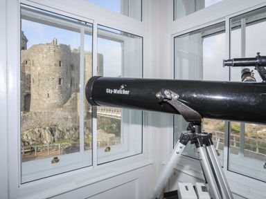 A black Sky-Watcher telescope on a tripod facing a window with a stone castle visible outside at Harlech Apartments - Branwen in Harlech