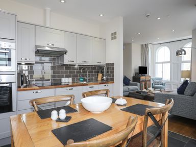 A kitchen and living area with a wooden dining table and chairs a gray tiled backsplash and gray cabinets at Harlech Apartments - Branwen in Harlech