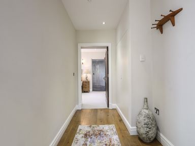 A hallway with wooden floor a rug a large decorative vase a coat rack and a view into a room with a bedside table lamp and a bathroom at Harlech Apartments - Branwen in Harlech