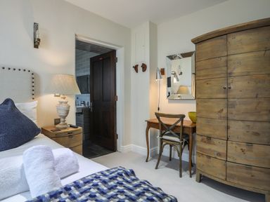 A bedroom with a bed with towels and a blue pillow a wooden nightstand with a lamp an open door leading to a bathroom a wooden desk with a chair a mirror and a large wooden wardrobe at Harlech Apartments - Branwen in Harlech