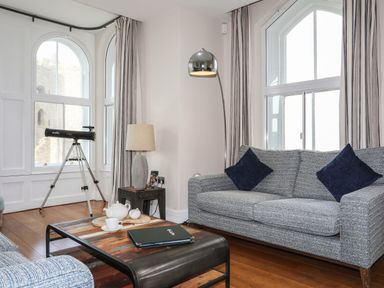 A sitting area with a gray sofa, a wooden coffee table with a teapot set, a telescope near large arched windows, and a floor lamp at Harlech Apartments - Branwen in Harlech