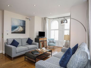 A living room with two grey sofas with blue cushions a coffee table a floor lamp an armchair a small cabinet with a TV and large windows at Harlech Apartments - Branwen in Harlech