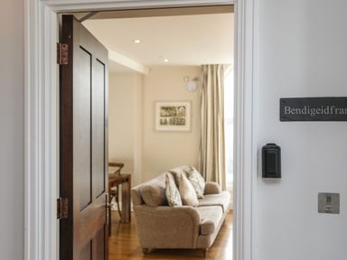 An open door revealing a living room with a beige sofa and cushions next to a window with curtains at Harlech Apartments - Bendigeidfran in Harlech