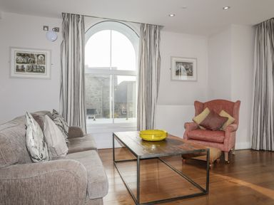 A living room with a sofa, armchair, wooden coffee table with yellow bowl, large arched window, and framed pictures on walls at Harlech Apartments - Bendigeidfran in Harlech