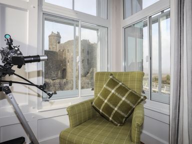 A green armchair with a matching cushion next to a telescope by windows showing a castle outside at Harlech Apartments - Bendigeidfran in Harlech