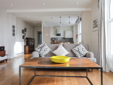 A living room with a grey sofa and three cushions a wooden coffee table with a yellow dish and an open kitchen in the background at Harlech Apartments - Bendigeidfran in Harlech